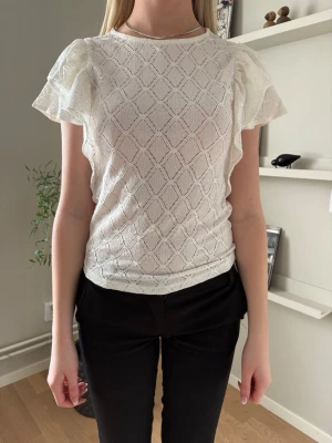 Vit spetsblus med volangärmar - Säljer en söt vit blus med spetsmönster och volangiga korta ärmar. Blusen har rund halsringning och ett diskret rutmönster i tyget. Perfekt att styla med både jeans och kjol för en trendig look.
