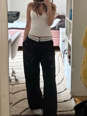 Svarta vida kostymbyxor med vit midjerand - Svarta kostymbyxor med bred passform och en snygg vit rand i midjan. De har medelhög midja och raka ben, vilket ger en avslappnad och trendig look. Perfekta för dig som gillar en chill och stilren vibe.