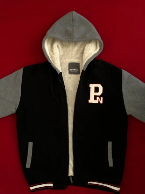 Svart och grå hoodie från Benhao - Snygg hoodie i collegemodell från Benhao med svart kropp och grå ärmar samt huva. Insidan är fodrad med mjuk, vit teddy. Broderad PN-logga på bröstet och randiga muddar i svart, vitt och rött. Två fickor framtill och dragkedja. Perfekt för en avslappnad streetwear-look.