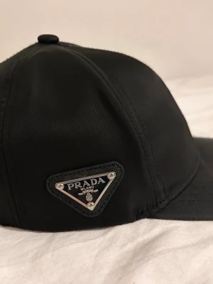 Svart keps från Prada med metallogo - Snygg svart keps från Prada med den ikoniska triangelformade metalloggan på sidan. Kepsen är tillverkad i ett glansigt, slitstarkt material och har klassisk böjd skärm. Perfekt accessoar för en trendig och stilren look.