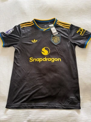 Manchester United matchtröja svart/gul - Snygg svart Manchester United matchtröja från Adidas med gula detaljer och logga. Tröjan har korta ärmar, klubbmärke på bröstet, sponsortryck och namn/nummer Mbeumo 19 på ryggen. Perfekt för fotbollsfans som vill sticka ut på läktaren eller planen.