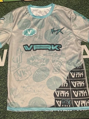 Grå VFRK t-shirt med turkosa detaljer - Säljer en grå t-shirt från VFRK med turkosa detaljer och tryck. T-shirten har korta ärmar, rund halsringning och är täckt av olika grafiska mönster och loggor i turkos och svart. Materialet känns lätt och andas, perfekt för en avslappnad stil.
