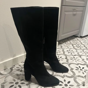 Svarta höga Calvin Klein mockastövlar - Säljer ett par svarta höga stövlar i mocka med bred klack och spetsig tå. Stövlarna har en stilren och trendig look som passar perfekt till höstens och vinterns outfits. Dragkedja på insidan för enkel påtagning. Nypris ca 2700, använda en kväll. Pris kan diskuteras vid snabb affär.