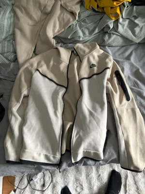 Beige nike tech fleece - Säljer ett par beige mjukisbyxor från Nike med dragsko i midjan och flera praktiska fickor, inklusive en med dragkedja. Byxorna har en avslappnad passform och är tillverkade i mjukt bomullsmaterial. Perfekta för chill dagar eller när du vill vara bekväm.