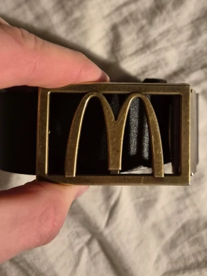 Svart bälte med guld McDonald's-spänne - Snyggt svart bälte med ett stort guldfärgat McDonald's-spänne i metall. Bältet är gjort i skinn och har en klassisk, stilren look med en unik twist tack vare det ikoniska M-loggan. Perfekt för dig som vill sticka ut lite extra.