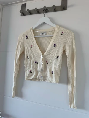 Beige kofta med broderade blommor JDY - Söt beige ribbad kofta från JDY med v-ringning och vita knappar framtill. Koftan har broderade små blommor i lila och rosa samt långa ärmar. Perfekt för dig som gillar en romantisk och trendig stil.