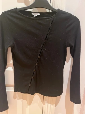 Svart långärmad topp med knappar - Snygg svart långärmad topp från amisu med dekorativa knappar i en diagonal linje framtill. Toppen har en rund halsringning och är tillverkad i ett ribbat material som ger en skön och stretchig passform. Perfekt att styla med jeans eller kjol.