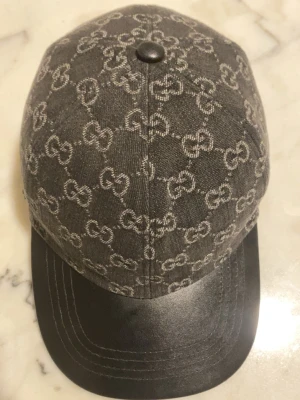 GG keps - Snygg keps från Gucci i grått tyg med klassiskt GG-monogram över hela kepsen. Skärmen är i svart läder och kepsen har en metallknapp på toppen. Perfekt accessoar för dig som vill ha en exklusiv och trendig look.
