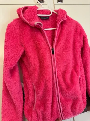 Rosa teddyjacka med huva - Supermjuk teddyjacka i starkt rosa färg från New Collection. Jackan har dragkedja framtill, två fickor och en mysig huva. Perfekt för dig som vill ha en varm och fluffig look. Långärmad och i fluffigt material som känns riktigt cozy.