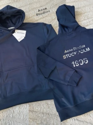 Mörkblå hoodie från Acne Studios - Snygg mörkblå hoodie från Acne Studios med klassisk huva och magficka. På ryggen står det 'Acne Studios STOCKHOLM 1996' i vitt tryck. Tillverkad i mjuk bomullsblandning och har ribbade muddar vid ärmslut och nederkant. Perfekt för en avslappnad men ändå fräsch look.