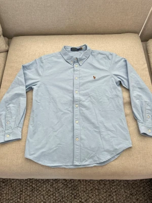 Ljusblå skjorta från Polo Ralph Lauren - Klassisk ljusblå skjorta från Polo Ralph Lauren med button-down krage och broderad logga på bröstet. Skjortan har långa ärmar och vita knappar framtill. Perfekt till jeans eller chinos för en clean och stilren look. Passar mer M 