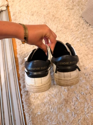 Givenchy vita sneakers med svart häl - Snygga vita sneakers från Givenchy med svart hälparti där det står 'Givenchy Paris' i guldtext. Klassisk låg modell med vita skosnören och robust sula. Tillverkade i läder för en lyxig känsla och stilren look.