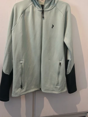 Ljusgrön hoodie med svarta detaljer - Snygg ljusgrön hoodie från Peak Performance med svarta muddar och svart huva. Dragkedja framtill och två fickor med dragkedja. Liten svart logga på bröstet. Perfekt för en chill och sportig stil.