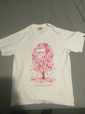 Vit bape Ape t-shirt  - Vit t-shirt från A Bathing Ape med ett stort rosa tryck av apans ansikte i form av blommor på framsidan och ett mindre tryck på ryggen. Klassisk passform och rund hals. Perfekt för dig som gillar streetwear och unika prints.