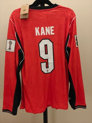 Spelar version England långärmad fotbollströja Kane - Röd långärmad spelar Version England fotbollströja från Nike med svart och vit detalj, nummer 9 och namnet Kane på ryggen. Tröjan har officiella FIFA World Cup-märken på ärmarna och Englands landslagslogga på bröstet. Perfekt för fotbollsfans!