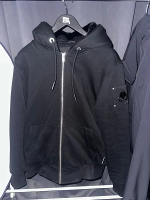 Svart hoodie från Moose Knuckles ÄKTA - Svart hoodie från Moose Knuckles med dragkedja, huva med dragsko och två stora fickor framtill. Mjuk, glansig insida och logotyp på ärmen. Perfekt för dig som gillar streetwear och vill ha något stilrent och bekvämt. Äkta kvitto finns vid förfrågan Köpt från Johnells.