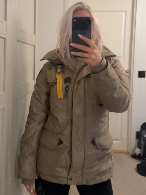 Beige jacka från Parajumpers - Säljer en beige dunjacka från Parajumpers i storlek S med gul rem vid kragen, flera fickor och patch på ärmen. Nypris ca 4500kr