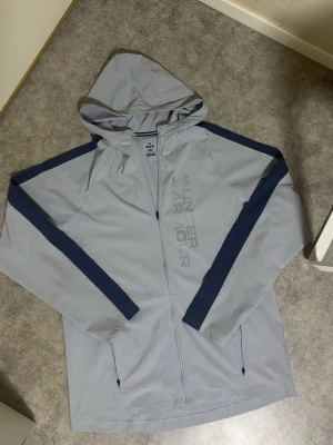 Grå vindjacka från Under Armour - Snygg grå vindjacka från Under Armour med marinblå ränder längs ärmarna. Jackan har huva, hel dragkedja och två fickor med dragkedja. Perfekt för blåsiga dagar och sportiga outfits. Stort Under Armour-tryck på bröstet och lätt, vattenavvisande material.