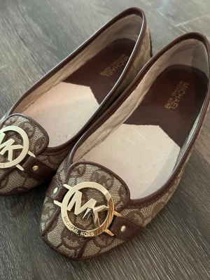 Bruna ballerinaskor Michael Kors - Snygga ballerinaskor från Michael Kors i brunt och beige med klassiskt MK-mönster. Skorna har rund tå, guldfärgad logga framtill och detaljer i läder. Yttersulan är svart och mönstrad för extra grepp. Perfekta för en trendig och stilren look. Fint skick.