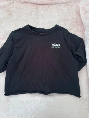 Svart croppad Vans t-shirt - Svart croppad t-shirt från Vans med liten logga och texten 'Off the Wall' på bröstet. Baksidan har ett stort tryck inspirerat av The Nightmare Before Christmas med en cool grafisk design. Perfekt för dig som gillar streetwear och unika prints.