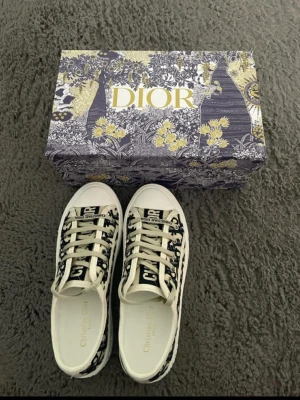 Dior Oblique sneakers i blått och vitt - Snygga Dior Oblique sneakers med blått och vitt monogrammönster, vita snören och robust vit sula. Klassisk låg modell med tydlig Dior-logga på sidan och tungan. Perfekta för dig som vill sticka ut med exklusiv streetstyle.
