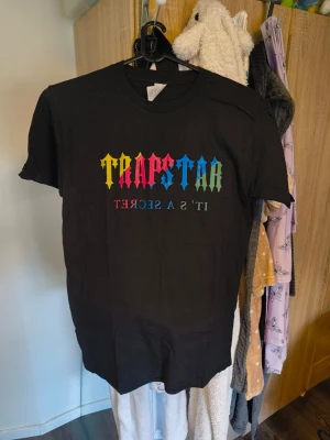 Trapstar svart set med färgglad text/m-l - Säljer ett svart set från Trapstar med t-shirt och shorts. Båda plaggen har färgglad Trapstar-logga och texten 'It’s a secret' tryckt på framsidan. Setet har en avslappnad passform och är gjort i mjukt material, perfekt för chill dagar.välj en storlek större finns m-xl Kan inte garantera äkthet 