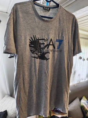 Grå EA7 Emporio Armani t-shirt/L - Snygg grå t-shirt från EA7 Emporio Armani med stort tryck av en örn och logga på bröstet. T-shirten har rund halsringning och korta ärmar. Perfekt för dig som gillar stilrena och sportiga plagg med en exklusiv touch. Kan inte garantera äkthet 