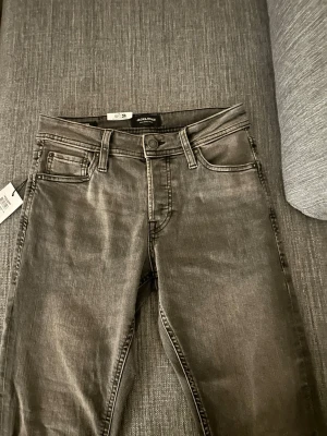 Grå jeans från Jack & Jones - Snygga grå jeans från Jack & Jones med klassisk femficksmodell och diskret slitning. Jeansen har normal midja, raka ben och coola detaljer på bakfickan. Perfekta för en avslappnad och trendig stil.