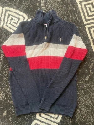 Randig stickad tröja från Polo Ralph Lauren - Mörkblå stickad tröja med dragkedja i halsen från Polo Ralph Lauren. Tröjan har breda ränder i vitt och rött över bröstet och ärmarna, samt broderad logga på bröstet. Ribbstickade muddar och hög krage ger en sportig vibe.