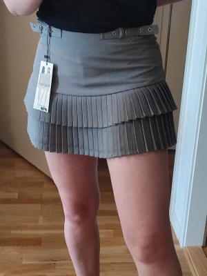 Y2K Trendig Mini Kjol - Super snygg grå kjol (skort, finns små shorts under). Storlek S. Helt nya med lapp kvar! Snygga detaljer i midjan i form av ett bälte som man kan dra åt.