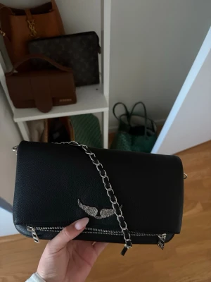 Svart Zadig & Voltaire handväska - Snygg svart handväska från Zadig & Voltaire i slätt läder med silverfärgad kedjerem och ikoniska vingdetaljen framtill. Väskan har dragkedjor och en stilren, rektangulär form som passar perfekt till en trendig look.