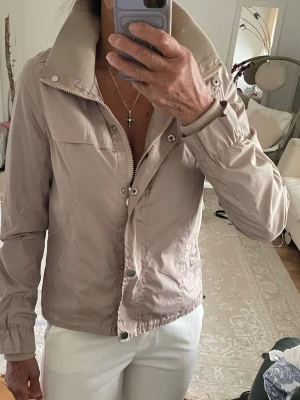 Beige vindjacka från Adrixx  - Snygg beige vindjacka från Adrixx med hög krage och dragkedja framtill. Jackan har långa ärmar med resår vid handlederna och praktiska fickor på sidorna. Perfekt för vår och höst när du vill ha en stilren look.
