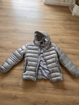 Grå pufferjacka från Polo Ralph Lauren - Snygg grå pufferjacka från Polo Ralph Lauren med huva och dragkedja. Jackan har två sidofickor med dragkedja och ikonisk röd logga broderad på bröstet. Perfekt för kalla vinterdagar och riktigt skön att bära tack vare det vadderade materialet.