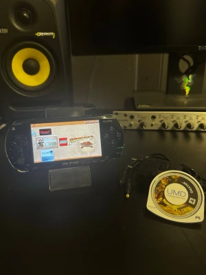 Sony PSP svart bärbar spelkonsol - Säljer en svart Sony PSP, en klassisk bärbar spelkonsol med stilren design i plast. Konsolen har en stor skärm, tydliga knappar och kommer med laddkabel samt ett UMD-spel. Perfekt för dig som gillar retrospel och vill ha gaming on-the-go.