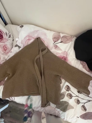 Beige stickad omlottkofta - Säljer en beige stickad omlottkofta med knytband i midjan. Koftan har lång ärm och är ribbstickad för en mysig och avslappnad look. Perfekt att slänga på sig över en t-shirt eller linne.