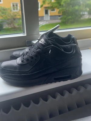 Nike Air Max 90 helsvarta sneakers - Säljer ett par helsvarta Nike Air Max 90 sneakers med klassisk siluett. Skorna har ovandel i läder och textil, synlig Air Max-dämpning i sulan och diskreta Nike-loggor på sidan och hälen. Perfekta för dig som gillar en clean och tidlös look.