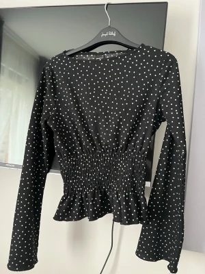 Svart prickig blus från Gina Tricot - Säljer en svart blus från Gina Tricot med vita prickar, långärmad modell och smockad midja som ger en snygg siluett. Blusen har rund halsringning och volangkant nedtill. Perfekt för dig som gillar trendiga detaljer och klassiskt mönster.