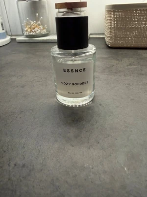 ESSNCE Cozy Goddess Eau de Parfum🤍🤍🤍 - Parfymflaska från ESSNCE med doften Cozy Goddess. Flaskan är genomskinlig i glas med svart cylinderformad kork och en enkel vit etikett. Modern och stilren design som passar perfekt på parfymhyllan.
