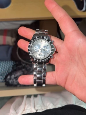 Silverfärgad BOSS herrklocka - Stilren herrklocka från BOSS med silverfärgad länk i metall och rund urtavla. Klockan har kronograf-funktion och tydliga markeringar runt boetten. Länken är robust och har BOSS-loggan ingraverad på spännet. Perfekt accessoar för en modern look.