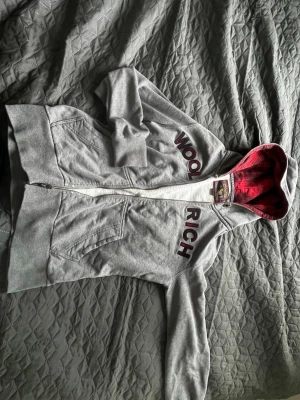 Grå zip hoodie från Woolrich - Snygg grå hoodie från Woolrich med dragkedja och stora fickor framtill. Märket står broderat i vinrött på bröstet och luvan har ett rött innerfoder i rutigt tyg. Perfekt för en avslappnad grisch-look. Tröjan är L men sitter som M