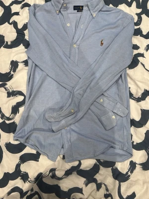 Ljusblå skjorta från Ralph Lauren - Klassisk ljusblå skjorta från Ralph Lauren med broderad logga på bröstet. Skjortan har knappar framtill och på kragen samt långa ärmar. Perfekt för en stilren och avslappnad look.