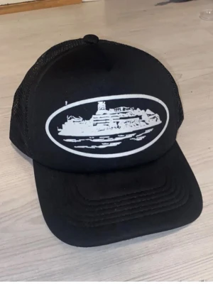 Corteiz keps - Säljer en svart truckerkeps med vitt tryck av ett skepp på framsidan. Kepsen har mesh på sidorna och justerbar snapback baktill. Perfekt för dig som gillar streetwear och vill sticka ut med en unik design.