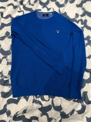 Blå stickad tröja från Gant - Snygg blå stickad tröja från Gant i premium bomull. Tröjan har rund halsringning, diskret struktur i materialet och Gant-logga broderad på bröstet. Perfekt för dig som vill ha en klassisk och stilren look.