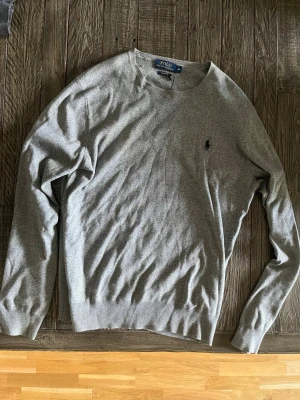 Grå tröja från Polo Ralph Lauren - Snygg grå tröja från Polo Ralph Lauren med rund halsringning och diskret broderad logga på bröstet. Tröjan har långa ärmar och ribbade muddar vid ärmslut och nederkant. Perfekt för en stilren och avslappnad look.