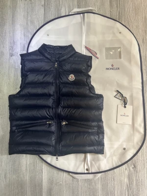 Moncler väst gui  - Hej! Jag säljer min navy blue gui väst. Den är i dunder skick med inga defekter eller liknande, säljs pågrund av att den inte passar min stil. Den är äkta och köpt på Nk. Allt i bilden ingår som dustbag och mer. Hör av dig vid frågor och liknande! 