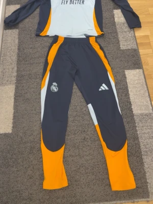 Real Madrid träningsset Adidas - Snyggt träningsset från Adidas och Real Madrid med långärmad tröja och byxor. Setet är mörkblått med orange och ljusblå detaljer, Real Madrid-logga och Adidas-märke. Perfekt för fotbollsträning eller sportiga dagar passar även m S xs men är S