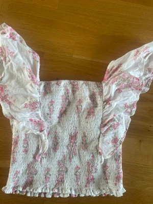 Blommig topp med volangärmar - Skriv innan köp! Supersöt topp i vitt med rosa blommigt mönster och volangiga, korta ärmar. Smockad kropp och små volanger längs nederkanten ger en romantisk vibe. Perfekt för dig som gillar en feminin och trendig stil. toppen är från Kappahl storlek 158/164! Pris kan diskuteras!🥰