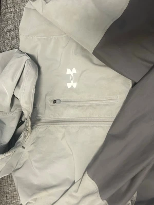 Grå och svart vindjacka Under Armour - Snygg vindjacka från Under Armour i grått och svart med huva och dragkedja på bröstet. Jackan har en sportig look, logga på bröstet och långa ärmar. Perfekt för blåsiga dagar och streetwear-stil. Pris kan diskuteras:)