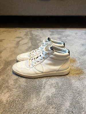 Vita high-top sneakers från Philippe Model - Snygga vita high-top sneakers från Philippe Model i läder med perforerade detaljer på tån och klassisk snörning. Skorna har en stilren design med diskreta paneler och en vit sula. Perfekta för dig som gillar en clean och modern look.