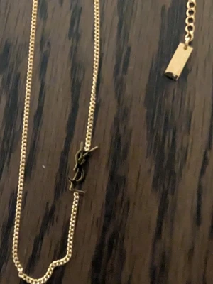 Guldhalsband med YSL-logga - Snyggt och stilrent halsband i guldton med den ikoniska YSL-loggan som hänge. Kedjan är tunn och har ett minimalistiskt rektangulärt avslut. Perfekt för dig som gillar exklusiva accessoarer med en lyxig touch.